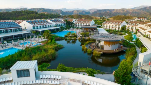 Dalaman Havalimanı'ndan Jiva Beach Resort oteline VIP ulaşım sağlayan lüks Mercedes Vito araç, arkaplanda Jiva Beach Resort lagün manzarası ve Çalış sahil şeridi.